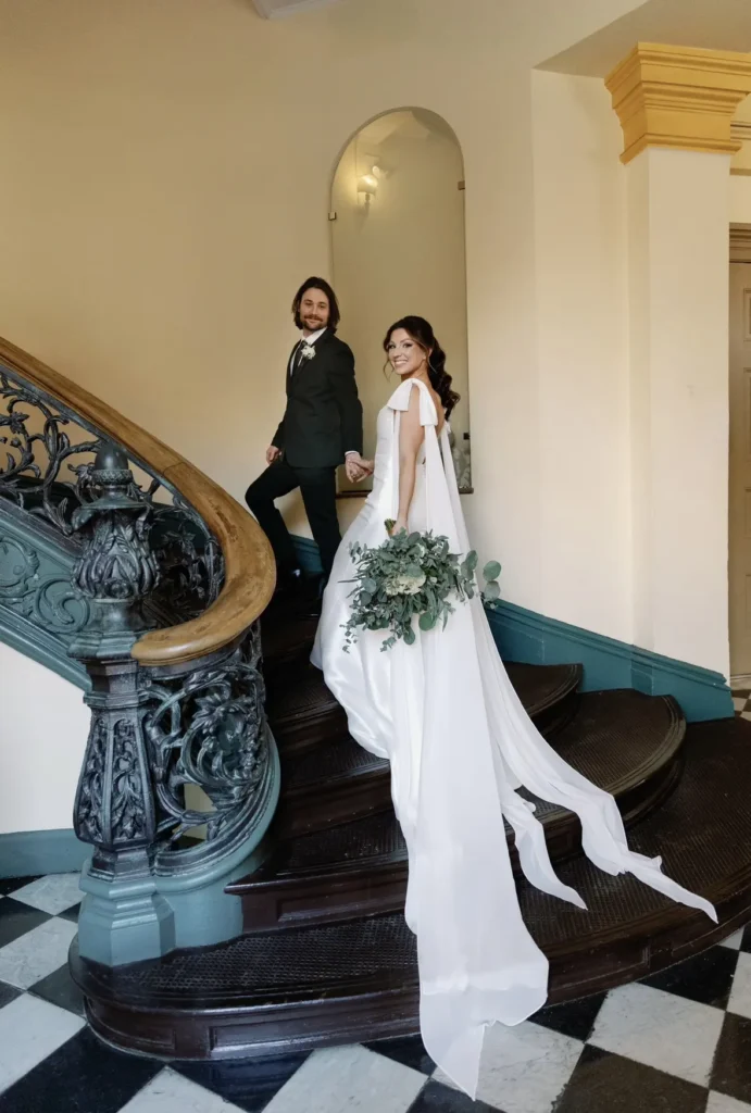 Dr. Sydney Nolan dressed in her wedding dress, ascending fancy stairs with her husband on their wedding day.